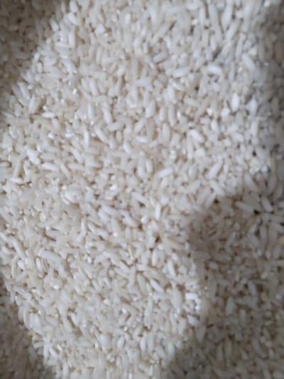 The image presents a close-up, top-down view of a large quantity of uncooked, white rice grains. The grains are primarily off-white to creamy in color and are spread across the entire frame, forming a dense, textured surface. Each grain is elongated and oval-shaped.

There are no people or discernible activities taking place. The scene focuses exclusively on the rice as the primary subject and object. Scattered among the white grains are a few tiny, dark specks, possibly indicating impurities or small fragments. The lighting appears even and diffused, not suggesting a specific time of day or outdoor environment. The depth of field is somewhat shallow, with parts of the rice in focus while other areas are softly blurred. No text or specific location cues beyond the rice itself are visible within the image frame. The provided location context is Gampongbaro, Indonesia.