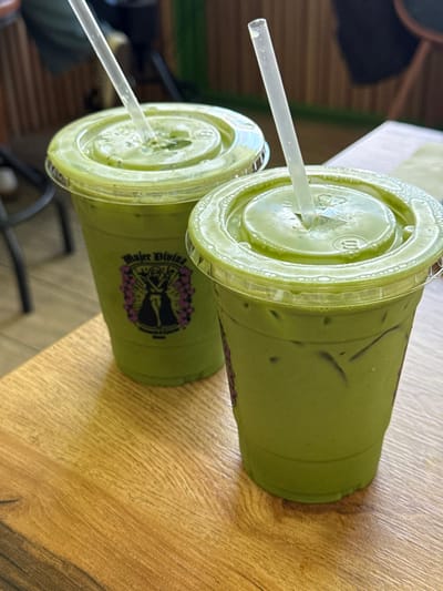 Two clear plastic cups containing iced light-green beverages are centered on a light brown wooden table with visible grain. Both cups are sealed with clear plastic dome lids and contain transparent plastic straws. The beverage in each cup contains ice cubes. The cup on the left features a label with the text "Mujer Divina" positioned above a dark silhouette figure framed by purple floral or wing-like designs, with smaller text "MATCHA & COFFEE" below. The background is out of focus, indicating an indoor setting, possibly a cafe or restaurant. The scene is located in Chula Vista, United States of America.