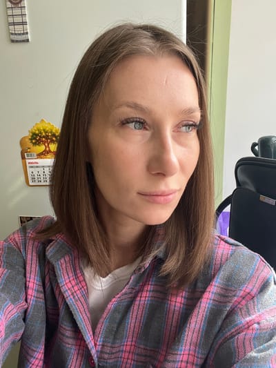 A young woman with shoulder-length brown hair is pictured in a medium shot, looking to her right. She is wearing a pink and gray plaid shirt over a white shirt. Her eyes are light blue and she has on mascara. Her skin is fair and smooth. A calendar showing the month of July is visible on the wall behind her, to her left. The calendar has an image of a tree with yellow lights on it. To her right, a black bag with a silver accent is visible. The background is a plain wall, part of which is painted green. There are no discernible landmarks to place this in Taldom, Russia.