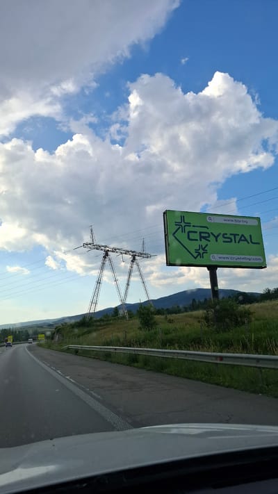 The image depicts a roadside scene on a clear day, with fluffy white clouds dotting a bright blue sky. A large, green billboard is prominently displayed on the right side of the frame. The billboard features the word "CRYSTAL" in large, bold, dark letters, along with a stylized logo of an arrow pointing left. Above and below the brand name, website addresses are visible: "www.bar.bg" and "www.crystalbgr.com". To the left of the billboard, a towering electricity pylon stands against the landscape, supporting multiple power lines that stretch across the scene. The pylon is characterized by its intricate metal lattice structure. The surrounding environment consists of rolling hills covered in green grass and scattered trees, with a mountain range visible in the background. A paved road with white lane markings runs diagonally across the lower left portion of the image, suggesting a highway or main road. Vehicles are present on the road, indicating ongoing traffic. The perspective is from a vehicle's interior, looking outward. The location is identified as Mirovo, Bulgaria.