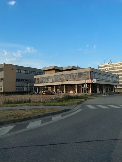 This is a photograph taken outdoors, likely in Strakonice, Czechia. The image is oriented vertically, with the sky occupying the left half and a street and building on the right. The sky is a bright, clear blue with a few wispy clouds.
The main subjects are a modern, multi-story building with a glass facade and a street with pedestrian crossing markings. The building appears to be an office building or commercial space. On the ground floor of the building, there is a sign that reads "RESTAURACE" which translates to "RESTAURANT." Above this, there is a round sign with a logo. The building has multiple floors with numerous windows. Next to the building is a paved road with a white pedestrian crossing. A grassy verge separates the road from the building.
There are no people visible in the photograph. The scene suggests it is daytime, likely late afternoon or early evening given the lighting. The weather appears to be clear and fair. There are no strong emotional cues conveyed by the image, it is a straightforward depiction of an urban or semi-urban setting. The overall impression is of a functional, possibly somewhat dated, piece of architecture next to a public thoroughfare. The presence of what appears to be construction materials or debris near the building's entrance suggests some ongoing work or a recent renovation.