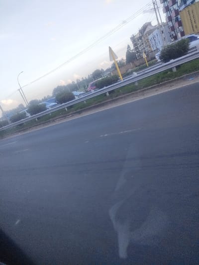 The image depicts a roadside scene in Murera ward, Kenya. The foreground consists of a dark asphalt road surface, appearing partially wet or reflective, with a faint, irregular white mark near the center-bottom. A metal guardrail separates the immediate road from a grassy median strip. Within this median, several uniformly trimmed bushes are visible, along with two yellow triangular warning signs mounted on yellow poles; one is prominent towards the right-center.

In the background, beyond the median, is another road or parking area containing multiple vehicles, including a light blue van-like vehicle and a red/pink passenger car. Further back, a line of dense trees and vegetation forms a horizon. To the right, a multi-story building with a light-colored facade, dark windows, and balconies is visible. Behind and to the right of this building, another taller structure appears, possibly under construction or featuring an unconventional design, with what resembles stacked shipping containers or modular units on its upper levels, some displaying yellow markings. Utility poles with overhead power lines crisscross the upper portion of the frame against a pale blue sky with scattered light clouds. No individuals are clearly discernible.