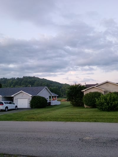 The image shows a suburban street scene with a row of houses on the right side and a road in the foreground. The houses appear to be single-story, with light-colored siding and dark roofs. There are well-maintained lawns in front of the houses, and trees and shrubs are visible. A white minivan is parked in the driveway of one of the houses. The sky is overcast with dramatic clouds. The scene appears to be in a residential area of Jeffersonville, United States, characterized by its hilly terrain covered in lush green trees.
