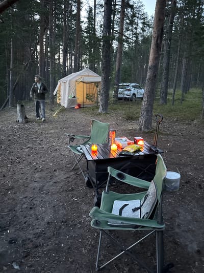 In Priladozhskoe, Russia, a campsite is set up in a pine forest. A light-colored canvas tent with glowing interior lights is visible in the background. A white SUV is parked behind the tent. In the foreground, a folding table is set up with two camping chairs. On the table are two lit candles, a package of snacks, and a phone. Another camping chair is positioned to the right of the table. A person wearing a green jacket and jeans walks in the distance, holding a cup. The ground is covered with pine needles and debris.