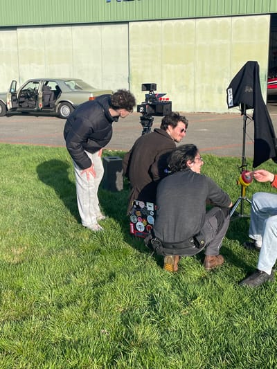 An outdoor scene at Chonas-l'Amballan, France, depicts four individuals engaged in what appears to be a video production against the backdrop of an airfield. The foreground is lush green grass under bright sunlight, casting distinct shadows. In the background, a light-beige hangar door and wall are visible, bordered by an asphalt tarmac with orange markings.
On the tarmac, a vintage light-beige Mercedes-Benz sedan (W126 S-Class) is parked with its front and rear passenger doors open. Adjacent to it, a professional video camera on a tripod is positioned, facing towards the right side of the frame. To the right of the camera setup, a C-stand holds a black fabric flag or silk.
Four male-presenting individuals are arranged around the camera and lighting equipment.
1. **Person 1:** On the far left, standing and leaning forward, wearing a black puffer jacket, light-colored pants, and light sneakers. He has curly brown hair and is looking towards the camera setup.
2. **Person 2:** In the middle-right, seated or crouching, wearing a dark brown coat and sunglasses. He has a dark beard and is looking towards the camera setup. A laptop covered in various stickers rests on the ground beside him.
3. **Person 3:** In the immediate foreground to the right of Person 2, crouching, wearing a dark grey long-sleeved shirt, dark utility pants with a belt, and brown boots. He has dark hair and glasses, and is looking towards the right with a smile.
4. **Person 4:** Partially visible on the far right, seated or crouching, wearing light blue jeans and black shoes. He is holding a small, round, red and yellow object in his right hand.
The individuals appear to be observing or working on the production, with direct sunlight indicating clear weather conditions.