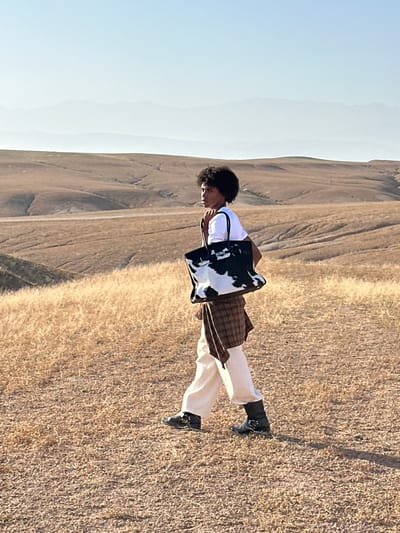 The image shows a person standing in a field with mountains in the background. The person is wearing a white t-shirt, light-colored pants, and black boots, and has a patterned tote bag. The location appears to be a dry, grassy area. While the image shares landscape characteristics with Sidi Badhaj, Morocco, there is no way to confirm that the scene is in that specific city from visual evidence alone. The person is facing to the left. The ground is covered in dry grass. The sky is clear and blue.