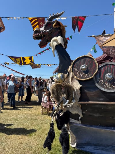 The image captures a vibrant outdoor event, likely a medieval fair, held under a clear blue sky. In the foreground, a section of a Viking ship's prow is visible, adorned with a fearsome dragon head figure with sharp teeth and red eyes. A skeletal figure dressed in a fur cloak and a helmet, carrying an axe, is positioned beside the ship. Two round shields are attached to the ship's side; one is labeled "HIDROMIEL" and the other "KOMB", presumably indicating mead and kombucha. Hanging from the ship are decorative black furry elements. In the background, a crowd of people are mingling, dressed in casual modern clothing, with some wearing hats. Colorful banners are strung across the sky, adding to the festive atmosphere. The ground is covered with dry grass, suggesting a warm climate. A tent with the word "COCTELES" is visible in the far left background. This scene is consistent with events like the "Mercado Medieval" held annually in the city of Llanera, Spain, known for its historical reenactments and cultural festivals.