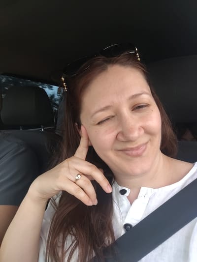 A woman with long, brown hair and light skin is sitting in the passenger seat of a car, looking at the camera and winking. She is wearing a white collared shirt with black buttons and a black seatbelt. She has a ring on her right ring finger and sunglasses on her head. The background shows a car interior with a dark headrest visible. The image does not provide enough information to determine the location in the city of Tselina, Russia.