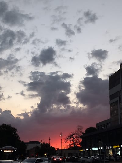 The image depicts a dramatic sunset over a city street scene. The sky is filled with dark, heavy clouds that are illuminated from below by a vibrant, deep red and orange glow. The clouds appear textured and voluminous, creating a sense of depth and drama.

In the foreground, a street is lined with parked cars and some buildings. The buildings have a modern, commercial appearance, with some showing illuminated signs. The silhouette of trees can be seen against the colorful sky. One building on the right is tall and modern with large windows. The streetlights and traffic lights are also visible, suggesting an urban environment.

The presence of vehicles and buildings indicates a city setting, and the text on one of the vehicles, "191 KLINIKA", suggests it might be a public transport vehicle. The overall impression is a visually striking sunset over a bustling city, with the natural beauty of the sky contrasting with the man-made structures. The image captures the fleeting moment of a colorful twilight.