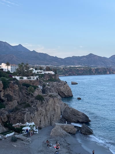 This is a vertical, outdoor, eye-level shot of a rocky coastline with a beach and the sea. The sky is a light blue, suggesting it is daytime, possibly late afternoon or early evening due to the soft light. The weather appears calm and pleasant.

In the foreground, large, textured rocks dominate the right side of the image, leading down to a sandy beach. A small, white structure with a trellis roof, possibly a beachside cafe or bar, is nestled amongst the rocks, with tables and chairs set up. A white umbrella provides shade.

Further down the beach, several people are visible. Some are lying on towels, sunbathing or relaxing, while others appear to be seated. Their clothing suggests casual beachwear. The water is a clear blue, with gentle waves lapping at the shore. A few smaller rocks are visible in the sea.

In the middle ground, a strip of white buildings with terracotta roofs is visible, dotted with palm trees and other greenery. This suggests a coastal town or resort. Behind the buildings, a steep, rocky mountainside rises, covered in sparse vegetation, and further back, larger, hazy mountains stretch into the distance.

The scene evokes a sense of relaxation and vacation. The natural beauty of the coastline is complemented by the human activity on the beach and the presence of the resort buildings. The lighting creates a peaceful atmosphere. No text is visible in the image.