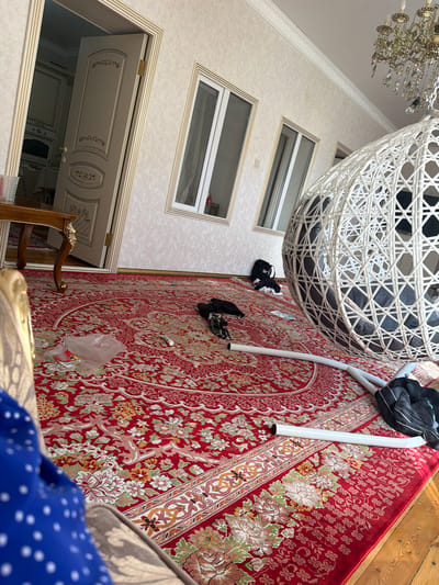 The image depicts an interior scene, likely a living room, in Khadjalmakhi, Russia. The room is dominated by a large, ornate red rug with intricate floral patterns. A white rattan hanging chair is suspended from the ceiling, with a dark cushion inside. Nearby, on the rug, are several dark-colored items that appear to be clothing or bags.

The setting includes a wall with textured wallpaper and several rectangular windows. Below these windows, a doorway with an open, cream-colored door reveals a glimpse of another room with decorative paneling. A grand, crystal chandelier hangs from the ceiling, suggesting a touch of elegance. The floor is partly covered by the rug and partly by wooden planks.

The time of day is not explicitly indicated, but the lighting suggests it is daytime. There is no visible information about the weather. The overall impression is of a well-decorated, perhaps slightly cluttered, residential space. The arrangement of objects on the rug might suggest a recent unpacking or a temporary placement of items. There is no visible text in the image.