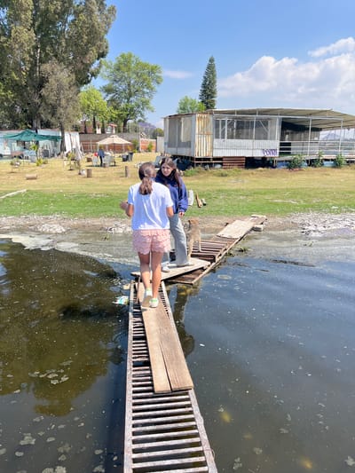 Two young females stand on a metal grate with wooden planks extending over a murky body of water in El Oasis Valsequillo, Mexico. The younger girl, facing away, wears a white shirt and pink lace shorts. The other girl, standing beside her, is older and wears a blue sweatshirt and grey pants, and looks toward the camera. The water is dark and speckled with light-colored debris. The shoreline is visible behind them, with a mix of grass, debris, and a concrete structure. Beyond the shoreline, there are trees and buildings. A blue sky with scattered clouds is above.