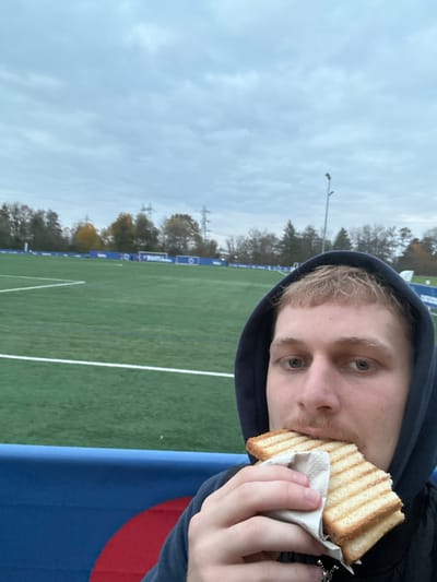 The image presents a close-up, selfie-style view of a male individual consuming a toasted sandwich at an outdoor sports facility. The scene is identified as a football (soccer) field, likely the Parc des Sports in Haguenau, France, evidenced by blue perimeter advertising banners, one partially legible as "PANTUM." The subject, a young adult with light hair, a light mustache, and blue eyes, is wearing a dark hooded sweatshirt with the hood up. He holds a grilled sandwich, characterized by distinct toast marks on the bread and partially wrapped in white paper, to his mouth.
In the background, a green artificial turf football field, marked with white lines, extends into the distance. Beyond the playing surface, a line of deciduous trees exhibits some autumnal foliage, while a tall metal utility pylon and a stadium light pole are visible against an overcast, cloudy sky. A blue barrier or railing is present in the immediate foreground, separating the subject from the field.