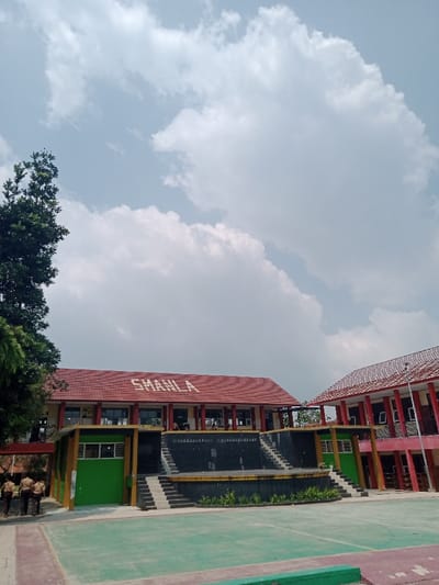 An outdoor wide shot depicts a school campus in Kawungluwuk, Indonesia, under a bright sky with scattered white clouds. The foreground features a light green paved court with white boundary lines. In the midground, a two-story building identified as "SMANLA" by white lettering on its red tiled roof dominates the left. This building has an open upper-level corridor with pillars and a lower level with windows, accented by green and yellow panels. A dark-colored, multi-tiered stage structure with steps is positioned directly in front of the SMANLA building. To the right, a second multi-story building with a red tiled roof and red/pink pillars is partially visible. Three individuals, dressed in brown uniforms, are standing on a concrete path in the lower-left corner, facing away from the camera. Faint figures are discernible on the upper corridor of the SMANLA building. A tall, leafy green tree partially frames the left side of the scene.