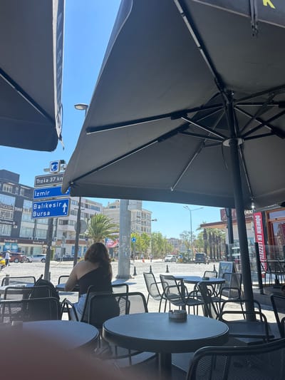 This image depicts an outdoor seating area at a cafe or restaurant, likely in Çanakkale Merkez, Turkey. The foreground is dominated by a large black umbrella, casting a shadow over the scene and suggesting it's a sunny day, possibly late morning or early afternoon. Several empty, dark metal tables and chairs are arranged on a paved area, with some featuring ashtrays.

In the background, a street scene unfolds. Traffic signs indicate directions to "Troia 37 km," "İzmir," "Balıkesir," and "Bursa," confirming the Turkish location. Buildings line the street, some with businesses advertising services, including one with a prominent red sign that reads "DÖNERİN EN LEZZETLI" (The most delicious döner). People are visible on the street and in the background, including a woman sitting at a table on the right side of the frame, facing away from the camera. The overall atmosphere appears relaxed, typical of an urban outdoor dining setting. The sky is a clear blue, reinforcing the impression of good weather.