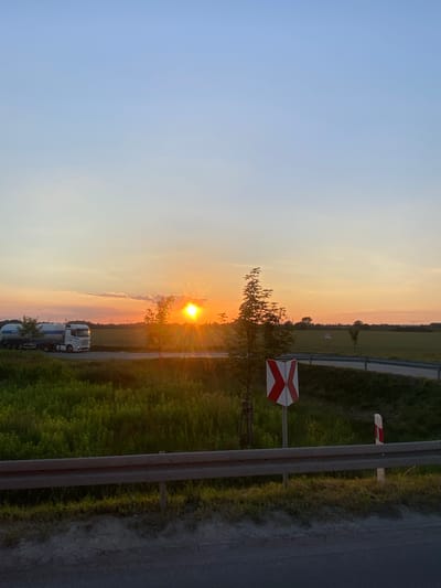 The image depicts a sunset scene in Podłęże, Poland. The sun is low on the horizon, casting a warm, orange glow across the sky and illuminating the landscape. The foreground features a road with a guardrail and a grassy verge. To the left of the road, a white and red striped sign, likely indicating a railway crossing, is visible. Further in the background, fields and trees stretch towards the horizon, with a few trucks parked on the side of the road. The sky transitions from a clear blue on the left to vibrant oranges and pinks near the sun, suggesting a beautiful evening. The overall mood is peaceful and serene, capturing the tranquil beauty of a rural sunset. No visible text is present in the image.