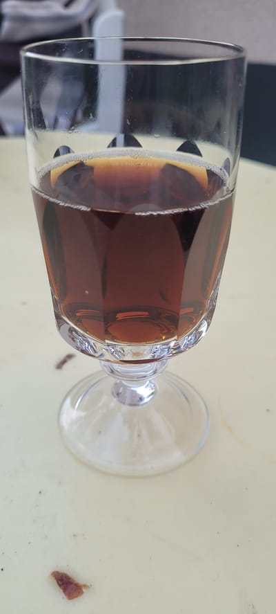 A close-up, eye-level shot shows a stemmed glass filled with a dark amber liquid, possibly iced tea or a similar beverage. The glass has a faceted design and rests on a clear, round base. The liquid has a thin layer of small bubbles near the rim. The glass is positioned on a light-colored, textured surface that appears to be a table. In the blurred background, a person in a chair is partially visible, suggesting an outdoor or semi-outdoor setting. A small, dark object, possibly food debris, is on the table in the foreground. The image does not provide any specific information about the city of Dietzenbach, Germany.
