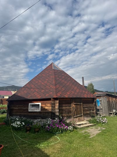 The image depicts a rustic wooden dwelling with a conical, shingled roof in Yabogan, Russia. The structure, likely a house or a shed, is made of weathered logs. A prominent feature is the reddish-brown shingle roof, which culminates in a small triangular window or skylight at its apex. The front of the building has a wooden door and a small, square window with decorative metal grilles.

In front of the dwelling, a colorful flower bed with white, pink, and purple blossoms adds a touch of vibrancy to the scene. Two decorative planters are placed within the flower bed. A small set of concrete steps leads to the doorway, and a path made of wooden planks forms a walkway across the green lawn.

To the right of the main structure, there is another wooden building with a sloped roof, appearing to be an attached shed or storage area. A blue plastic bag is visible hanging on its exterior. In the background, a metal chimney protrudes from the roof of the main dwelling, and power lines stretch across the sky. Further in the distance, rolling hills with some trees and other buildings can be seen under a partly cloudy sky. The overall impression is that of a rural, perhaps summertime, setting.