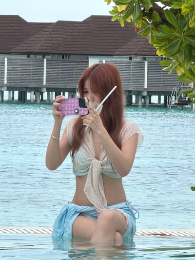 A woman is photographed in the Maldives, specifically in Guraidhoo. She is standing in a pool of water, holding a phone with a pink case and taking a picture. She has reddish-brown hair and is wearing a light-colored top and blue shorts. The pool is next to a wooden structure over the water. The water is calm, and there are some trees in the background.