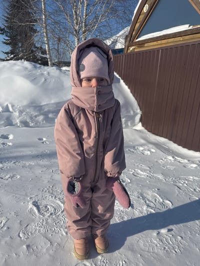 A young child stands in a snow-covered outdoor environment, identified as Устиново, Russia. The child, facing forward, is dressed in a full-body, mauve-pink snowsuit with a hood, a matching knit hat, and pink mittens. Only the child's blue eyes and the bridge of their nose are visible; the mouth and lower face are concealed by the snowsuit's high, snapped collar. The child wears light brown, puffy winter boots. The ground is covered in undisturbed white snow, with some faint footprints visible. Overhead, the sky is clear and blue, indicating bright daylight, and shadows are cast on the snow from the child. In the background, to the left, are snowdrifts and bare deciduous trees, possibly birches, along with a dark green evergreen tree. To the right, a brown corrugated metal fence runs horizontally, partially obscuring a light-colored wooden structure with a triangular attic window.