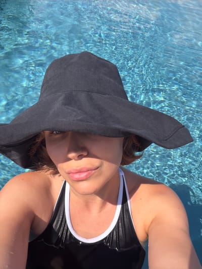 A woman wearing a black floppy hat and a black and white swimsuit is submerged in the clear blue water of a swimming pool. The sunlight reflects off the water, creating a shimmering effect. The woman's face is partially obscured by the brim of her hat, but her lips and chin are visible. Her eyes are looking towards the camera. The background is entirely composed of the rippling water of the swimming pool. The location is not specified as being in the city of Asciano, Italy.