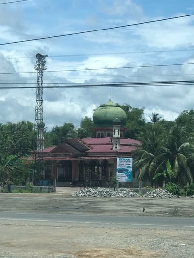 The image shows a mosque in Bulo Gogo, Indonesia, with a green dome and red roof. The mosque is situated next to a road. A communication tower is visible to the left of the mosque. Overhead power lines stretch across the sky, which is cloudy with patches of blue. A billboard is attached to the side of the mosque. Trees and other vegetation are present in the background. The ground is a mixture of pavement and gravel.