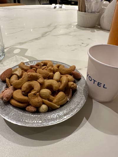 The image depicts an indoor scene on a light-colored, veined countertop, likely in a establishment such as a hotel lounge or bar in Zeytinburnu, Turkey. In the foreground, a round, silver-toned metal bowl with an embossed decorative pattern holds a mix of roasted nuts, including cashews, almonds, and peanuts. To the right of the bowl, a white paper cup displays the partial blue lettering "VOTEL", consistent with the Novotel brand. Directly behind the cup, an orange-colored bottle or container is partially visible. In the out-of-focus background, additional white ceramic containers are present, one holding multiple thin, light-colored sticks, possibly stirrers or toothpicks. No individuals or explicit actions are depicted.