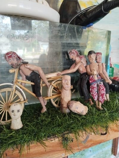 This image presents a close-up view of a miniature diorama, likely a cultural craft display from Jetis, Indonesia. The central focus is a group of four handcrafted figurines, appearing to be made of wood or clay, depicted in the act of pushing a two-wheeled wooden cart or bicycle.
One figure is positioned at the front of the cart, leaning forward and holding its structure, with a red patterned bandana on his head and dark trousers. Immediately behind him, another figure also pushes the cart's frame, wearing similar attire. Two more figures stand to the right, behind the cart. One of these wears distinctive red and white patterned shorts or a sarong and holds a small, carved stick-like object in his hand, while the fourth figure is partially obscured. All visible figures have head coverings. Their postures convey effort or struggle.
The miniature cart itself is intricately detailed, with wooden wheels and spokes. The figurines are placed on a vibrant green artificial grass mat, which rests on a natural wood plank surface. In front of the cart, on the grass, are two detached carved wooden heads or busts; one is upright, and the other lies on its side.
Behind the entire display is a clear, somewhat dirty or aged glass enclosure, possibly a display case or an old aquarium. Through this glass, blurred background objects are visible, including a prominent white plastic object on the left and a black appliance, possibly a hairdryer, on the right. Faint, blurry text, appearing to start with "Dongkrak," can be discerned through the glass on the left side, though it's largely illegible. The scene depicts a narrative of manual labor or a journey.