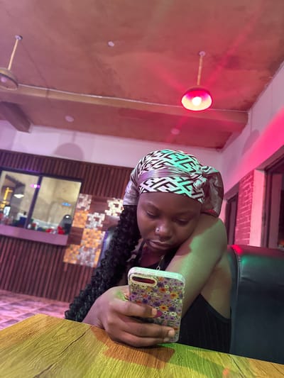 In Laminga, Nigeria, a young woman is seated at a wooden table, engrossed in her mobile phone. She wears a headscarf with a geometric pattern and has long, dark braids. The environment appears to be an indoor establishment with exposed ceiling beams, punctuated by pendant lights casting red and green hues. The background features a wall with vertical wooden slats and a patterned mosaic, and a large window or partition reveals more of the interior. The lighting creates a vibrant, somewhat moody atmosphere.