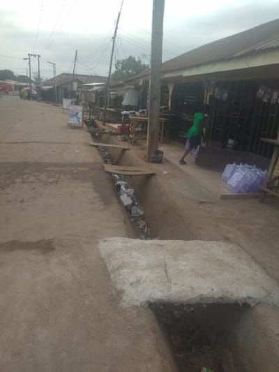 The image shows a dirt road in Junction, Nigeria, with a drainage ditch running down the middle. The ditch is partially covered by a concrete slab. There are buildings on the right side of the road, some with barred windows and a sign that reads "Rex Paints". A single person is walking towards the right side of the image, carrying bags of what appears to be water in plastic bags. The road is relatively empty except for a motorcycle parked near the left side.  Power lines run across the image. The sky is gray and overcast.  The ground is dusty and dry.