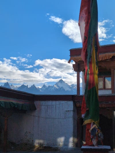 A prayer flag flutters in the foreground, with a building and snow-capped mountains in the background under a blue sky with clouds. The building appears to be a monastery or temple, with traditional architecture featuring wooden beams and decorative trim. The mountains are rugged and imposing, with snow covering their peaks. The prayer flag is made of multiple colored panels, likely representing the five elements of Buddhism. The overall scene suggests a peaceful and spiritual setting in a mountainous region. The location is identified as Yulang, India, a place known for its Buddhist monasteries and stunning natural beauty.