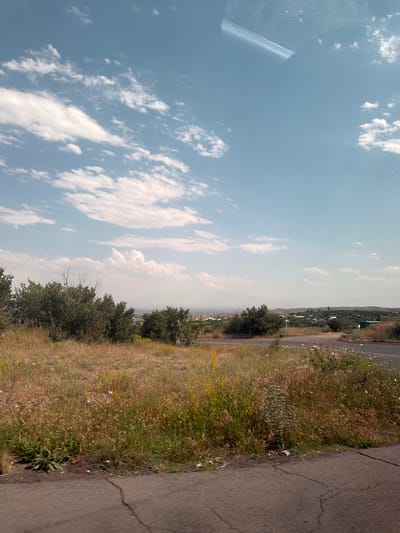 The image depicts a rural landscape with a road in the foreground and a village in the middle ground. The sky is partly cloudy with white clouds scattered across a blue expanse. The foreground is dominated by dry grass and some green bushes, suggesting a warm climate. The middle ground features a village with houses, nestled amongst green trees and rolling hills. In the distance, mountains are visible, partly obscured by haze. The road appears to be asphalt and has a faint white line marking the edge. There are no people or animals visible in the scene. The overall impression is of a serene, undeveloped area. The location is specified as Byurakan, Armenia.
