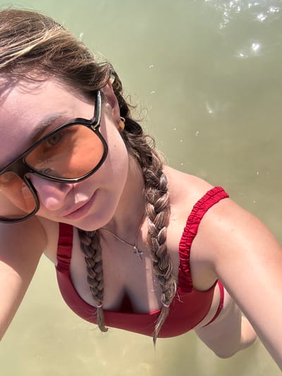 A young woman with braided hair and sunglasses poses for a selfie in shallow, murky water. She is wearing a red bikini. Her face is tilted downwards, and she is smiling. The water is a light brown color, and there are small ripples on the surface. The background is blurry and out of focus.