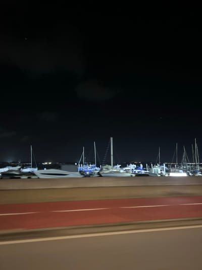 A nighttime view features numerous boats with masts aligned along a dark body of water, possibly a harbor or dock. The masts are illuminated by lights. The view is partially framed by a window structure consisting of a beige and red border. The contrast between the dark water and the illuminated boats creates a strong visual.