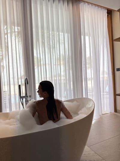A woman with wet, dark hair is relaxing in a white, oval-shaped bathtub filled with bubbles. Her back and part of her shoulder are visible above the foam. The tub is positioned in front of a large window covered with sheer, white blinds. The overall scene appears to be a luxurious bathroom setting with an emphasis on relaxation and privacy.