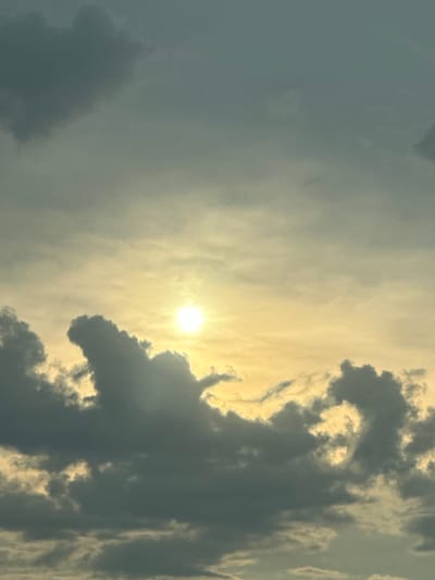 The image shows a sky with a bright sun peeking through dark, dramatic clouds. The sun is located in the upper center of the frame, casting a warm yellow and orange glow. The clouds are a deep grey, with some illuminated edges where the sunlight catches them. The overall impression is one of a dynamic and somewhat moody sky. There are no visible people, structures, or any recognizable landmarks that would indicate a specific location within the city of Skorotovo, Russia. The scene is purely atmospheric.