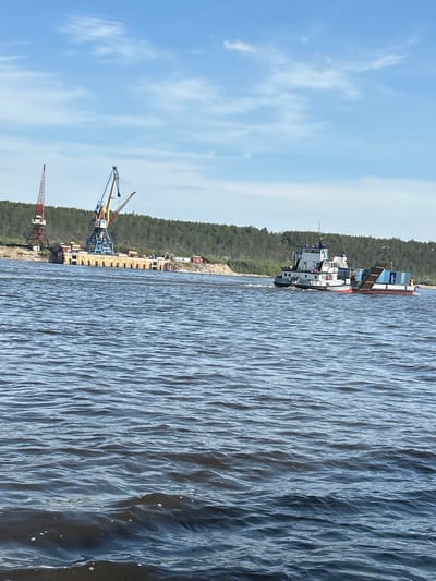 The image depicts a wide, dark blue river in Nizhny Bestyakh, Russia, under a partly cloudy sky. In the background, a forested hill forms the horizon. On the riverbank, a dock with several cranes is visible, including a red and white tower crane on the left and a blue and yellow gantry crane in the center. A tugboat is moving across the river, towing a barge laden with several blue containers. The water is choppy with small waves.