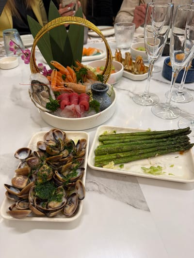The image shows a table spread with food, including a platter of sashimi with oysters and shrimp, a dish of clams with a green herb topping, and a plate of asparagus. Champagne flutes are visible in the background, along with a small vase with a pink flower. The setting appears to be a restaurant or dining establishment. There is no indication that this is in the city of Torreforta, Spain. People's hands are seen near the food. The tabletop has a white marble-like appearance.
