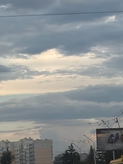 The image captures a cloudy sky over a cityscape, likely Shcherbani, Ukraine, based on the provided context. In the foreground, a multi-story apartment building with beige and white facades is visible on the left. Power lines and utility poles crisscross the sky, suggesting an urban environment. To the right, a billboard displays an advertisement for a car, with the text "Новий Tayron" (New Tayron) partially visible. The sky is filled with a mix of dark gray and lighter, possibly overcast, clouds, with some hints of pale yellow light breaking through. Evergreen trees are scattered throughout the scene, some near the apartment building and others in the middle ground. The overall impression is one of a typical urban landscape under a cloudy sky.