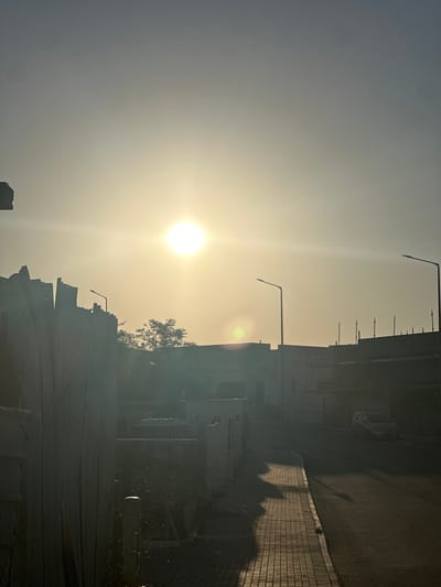 The image captures a bright, hazy sun low in the sky, casting a warm, yellowish light over an urban scene in Arad, Israel. The foreground features a textured, dark vertical structure, possibly a fence or wall covered in netting. To the right, a paved sidewalk runs alongside an asphalt road, with the sun creating long, distinct shadows stretching across the pavement. A few sparse plants grow along the edge of the sidewalk. In the mid-ground, several buildings are visible, some appearing to be under construction with scaffolding. Streetlights stand tall along the road. A car is parked on the right side of the road. The background is dominated by the sun and the hazy sky, with silhouettes of trees and more buildings. The overall impression is of an outdoor environment, likely a street or construction area, bathed in the soft, diffused light of a sunrise or sunset.