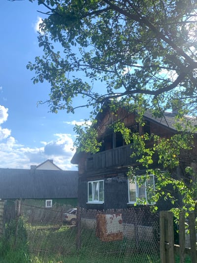 An outdoor scene in Yasinia, Ukraine, depicts a residential area under clear blue skies with scattered white clouds. In the foreground, a dark, rusted chain-link fence with a wooden post on the right spans the lower frame. Overhanging tree branches with green leaves partially obscure the upper and right portions of the view.

The midground features two structures. On the right, a two-story wooden house with dark-stained paneling and a dark roof stands prominently. Its ground floor contains two rectangular windows with white frames. The second story includes an open wooden balcony. Faint disrepair is visible on the facade below the balcony. Attached to the fence in front of this house is a faded, partially obscured rectangular sign with red and white elements. To the left, a long, single-story building painted green is visible behind the fence, featuring a dark, ridged roof and one small white-framed window. A light-colored vehicle is partially visible behind the fence and in front of the green building.

In the background, the light-colored gable and dark roof of another building are visible beyond the green structure. Bright sunlight illuminates the scene, creating highlights on foliage and reflecting off surfaces. No individuals are present.