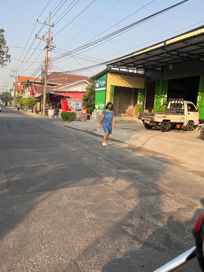 The image captures a scene on a street in Sugih Waras, Indonesia, bathed in the warm light of what appears to be late afternoon or early morning. A woman wearing a blue dress and a green face mask walks on the asphalt road. She has short, dark hair and is wearing white sandals. She is holding a clear plastic bag in her right hand.
To her right, a white pickup truck with a black roll cage is parked on a paved area next to a building with a yellow and gray awning. The building has green folding doors, and stacks of what appear to be building materials or cement bags are visible inside and outside. A sign with the text "Avian BRANDS" is prominently displayed on the building.
The road itself is asphalt with visible patching and wear. Trees line the street, and in the background, residential buildings with red roofs can be seen. Utility poles with a complex network of wires stretch across the sky. The overall atmosphere suggests a typical street scene in a commercial or mixed-use area of the town.