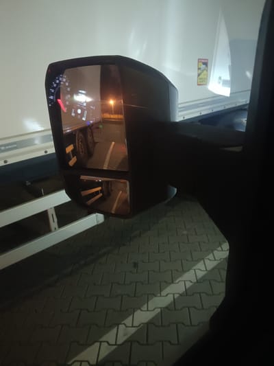 This image shows a view from the driver's side of a truck, looking into the side mirror. The side mirror reflects the trailer of the truck, a speed gauge on the dashboard, and a street light. The trailer is white with some reflective markings and a red and yellow sticker on the side. The speed gauge shows a reading between 0 and 200, with the needle pointing to approximately 40. The background in the mirror appears to be a dark, possibly paved area, with a street light illuminating the scene. The overall setting suggests that the truck is parked or stopped at night. The image was taken in Tyniec Mały, Poland, as indicated by the metadata.