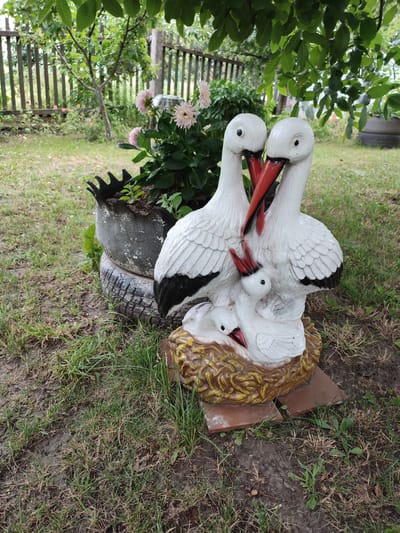 In Voskobiinyky, Ukraine, a garden scene features a white ceramic sculpture of storks family placed in a nest. The sculpture depicts two adult storks and two chicks, nestled together. The adult storks have red beaks and black wing tips. Behind the sculpture, a wooden fence encloses a garden with green grass and a bush with pink flowers. The sculpture is positioned on a base of broken terracotta tiles. The surrounding environment includes dirt paths and dry grass, suggesting a natural, outdoor setting.