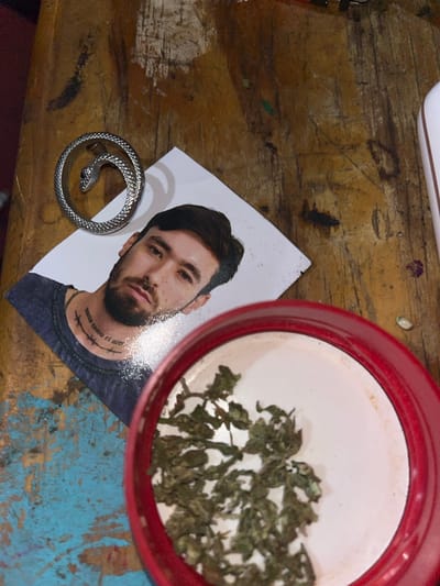 A photograph taken at an undisclosed location in Llavallol, Argentina, depicts a tabletop arrangement. A man's photograph is positioned to the left. Adjacent to the photograph is a silver ring shaped like a snake. To the right, a red container holds what appears to be dried herbs or plant matter. The surface beneath these items is distressed, with visible wear and tear.