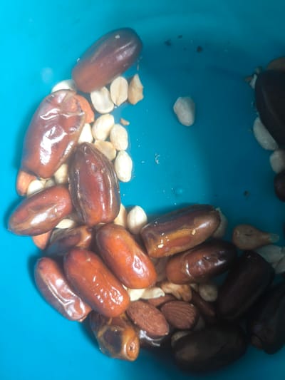 This media file shows a close-up, overhead view of a bright blue bowl filled with a mixture of dates and nuts. The dates are various shades of brown, from lighter reddish-brown to very dark brown, and appear to be dried. Some of the nuts are lighter brown and almond-shaped, while others are off-white and oval, resembling peanuts or macadamia nuts.

The subjects are the food items within the bowl. The setting is clearly a bowl, likely a food preparation or serving bowl, given its contents. The lighting suggests it is daytime, possibly indoors or in a shaded outdoor area, as there are bright highlights on the smooth surfaces of the dates, indicating a light source. There are no people, text, or specific location cues to determine the time of day or weather. The overall impression is that of a healthy snack or ingredient mix. The bowl is filled, with the food items occupying most of the frame, suggesting a generous portion.