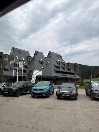 In Žunovnica, Bosnia and Herzegovina, a multi-story hotel with a distinctive, angular, dark-grey roof is visible. The building's facade incorporates wood paneling and large glass sections. Two flags are mounted on flagpoles in front of the hotel, one appearing to be the flag of Bosnia and Herzegovina. A parking lot is in the foreground, with several vehicles parked. These include a black Mercedes-Benz SUV, a teal Volkswagen T-Cross, a black Mercedes-Benz sedan, and a grey SUV on the far right. The weather appears overcast. A forest of pine trees borders the area behind the hotel.