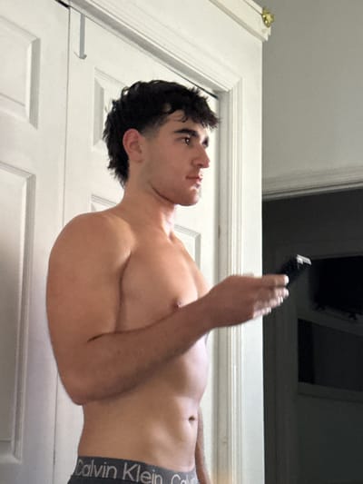 A young man, shirtless and wearing Calvin Klein underwear, is standing indoors, holding a black electric trimmer in his right hand. He is looking to his right, with his hair styled in a modern mullet. The background consists of a white doorframe with paneling, and a dark doorway leading to another room where a television is visible. The image appears to be a personal photograph taken in a domestic setting in Bellmore, United States of America.
