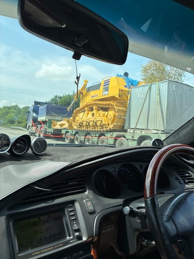 The image shows a view from inside a vehicle, looking forward through the windshield. The vehicle is on a road, and in front of it, a large yellow Komatsu bulldozer is being transported on a flatbed truck. The bulldozer is labeled "275A". Adjacent to the bulldozer on the truck is a large, corrugated metal container. A blue tarp covers part of another truck parked behind the flatbed. The surroundings appear to be rural or semi-rural, with trees visible in the background and a cloudy sky overhead. The interior of the vehicle shows part of the dashboard with gauges and a navigation system. The image was taken in Spassk-Dal'nij, Russia.