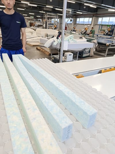 In a Zangiata, Uzbekistan manufacturing facility, a man in a dark blue t-shirt with a "FoamLine" logo and blue pants stands with his hands in his pockets. In the foreground, four large, rectangular foam blocks are arranged diagonally on a white surface covered with a pattern of small, white, oval-shaped objects. The foam blocks are a mix of white and light blue colors, with some flecks of pink and yellow visible. The environment appears to be a production line, with various machinery and stacks of materials in the background, suggesting a factory setting.